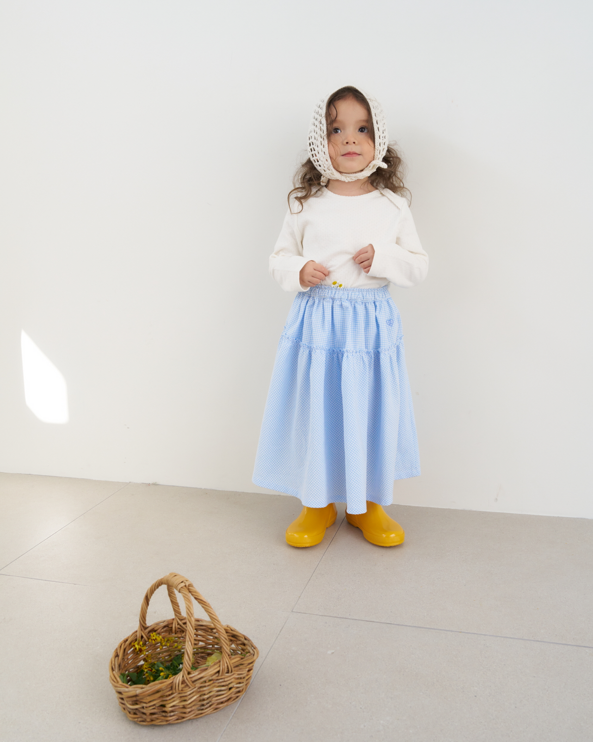 Child wearing The Fruto Clair Check Skirt in Blue Check, a light blue tiered gingham skirt styled with a white top, knit bonnet, and yellow rain boots.